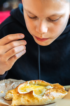 Boy eating traditional khachapuri bread with cheese and egg filling on wooden board close-up view, concept of traditional cuisine enjoymentの写真素材