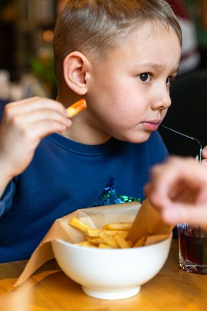 Young boy eating French fries and sipping a drink through a straw at a restaurant table close-up view, concept of casual diningの写真素材