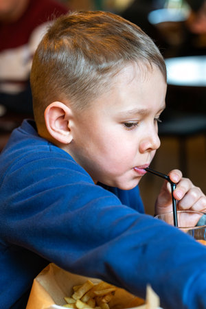 Young boy eating French fries and sipping a drink through a straw at a restaurant table close-up view, concept of casual diningの写真素材