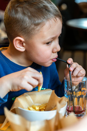 Young boy eating French fries and sipping a drink through a straw at a restaurant table close-up view, concept of casual diningの写真素材