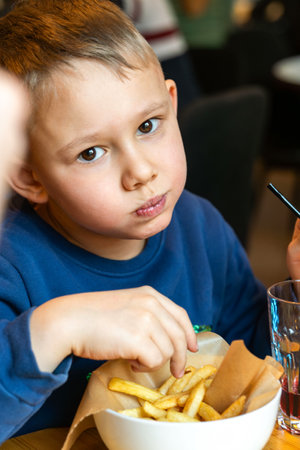 Young boy is eating French fries and drinking soda at a restaurant table indoors close-up, concept of casual dining funの写真素材