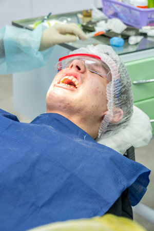 Young patient with his mouth open lies in a dental chair while a professional performs treatment in a well-lit clinic, wearing protective gearの写真素材