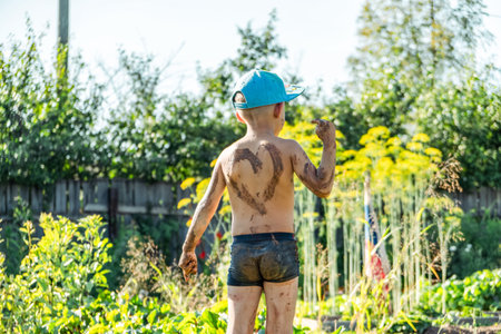 Little boy wearing a cap stands in a garden, covered in mud and proudly displaying a heart shape on...