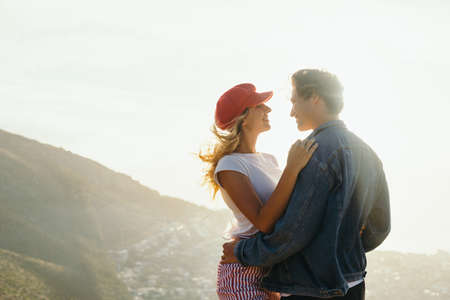 Happy romantic caucasian young couple smiling and looking at each other while embracing on mountain against sky during sunset at weekendの写真素材