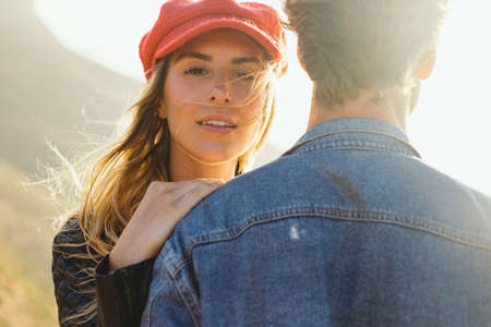Close-up portrait of beautiful young woman wearing cap with hand on boyfriend's shoulder enjoying sunset with him during weekend outingの写真素材