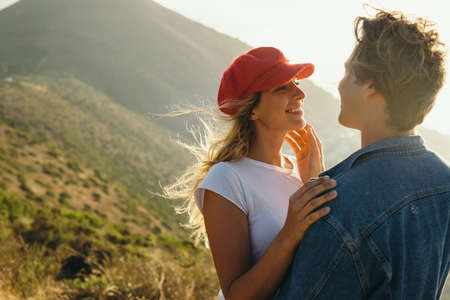 Romantic caucasian young boyfriend touching beautiful smiling girlfriend's face while enjoying weekend on mountain during sunsetの写真素材