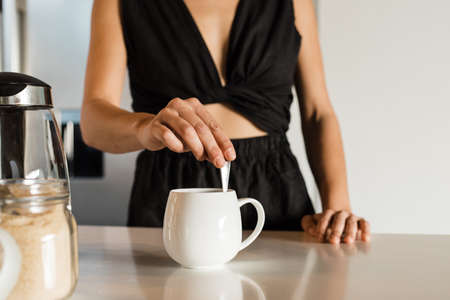Midsection of mixed race mid adult woman wearing black attire mixing coffee in cup on kitchen island in morningの写真素材