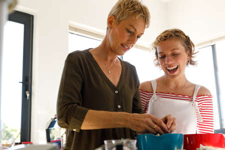 Low angle view of excited teenage daughter with mouth open laughing and looking at mother preparing...