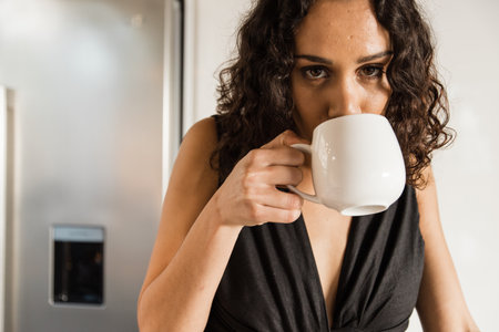Portrait of mixed race beautiful mid adult woman with curly hair drinking fresh coffee in kitchen during morningの写真素材