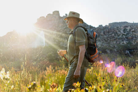 Side view of mature man wearing hat with backpack hiking amidst plants against sunlight streaming through mountain at sunsetの写真素材