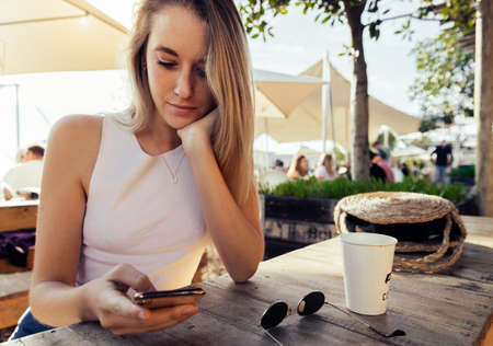 Beautiful blond woman with hand on chin using mobile phone while sitting by coffee cup and sunglasses at wooden tableの写真素材