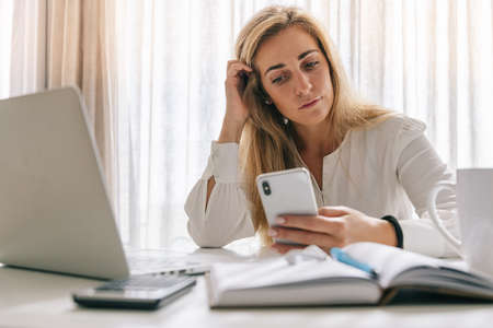 Mid adult blond female freelancer with diary, laptop and calculator on desk using smartphone while working in home officeの写真素材