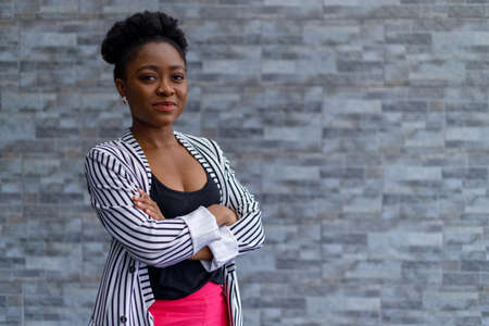 Portrait of african american confident millennial businesswoman with arms crossed standing against brick wall outside officeの写真素材