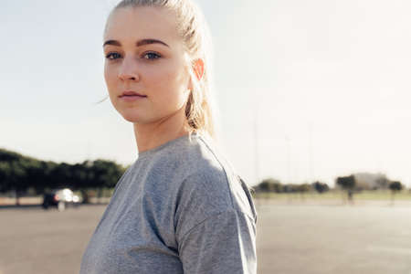 Portrait of confident young female athlete with blond hair looking at camera while standing on street against clear skyの写真素材