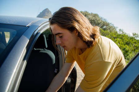 Young male traveler with long hair standing by car while enjoying vacation during sunny dayの写真素材