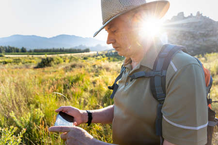 Sunlight streaming behind mature male explorer wearing hat using mobile phone while hiking on lush...