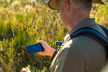 Mature male backpacker using mobile phone while hiking on lush grassy land at countryside during...