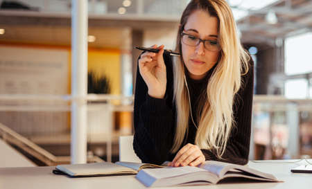 Focused young caucasian woman with blond hair wearing eyeglasses and studying at desk in library at...
