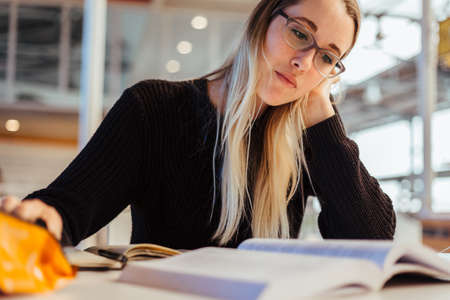 Focused young caucasian woman with blond hair wearing eyeglasses and studying at desk in library at...
