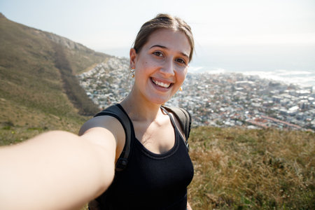 Portrait of smiling young female explorer with backpack standing on mountain with cityscape, sea and clear sky in background during summerの写真素材