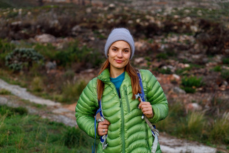 Portrait of smiling female caucasian hiker in winter clothes standing in front of mountain trail in...
