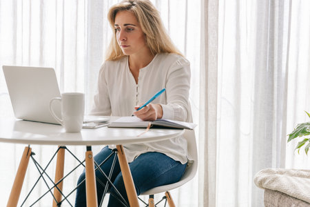 Smiling mid adult female freelancer with laptop on table writing notes in diary while working in home officeの写真素材