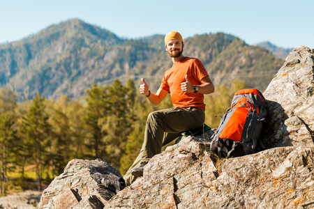 A young male with a beard is traveling through the mountain, a tourist with a rucksack standing on the rock hill while enjoying nature view, summer holidays in the mountains.の写真素材