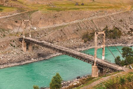 Old ruined wooden bridge over the turquoise river in the mountains in cloudy weatherの写真素材