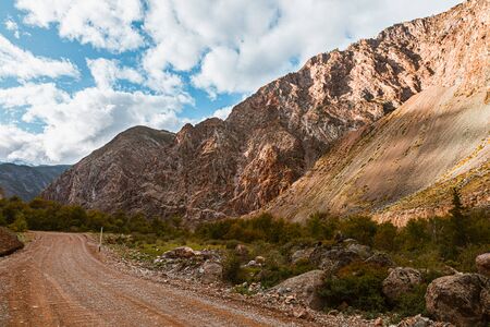 Panorama of road to the gorge of mountains on background of beautiful snowy mountain peaks in cloudy weather. Mountain road in the forest. View of trail on the background of blue sky.の写真素材