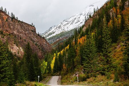 Panorama of road to the gorge of mountains on background of beautiful snowy mountain peaks in cloudy weather. Mountain road in the forest. View of trail on the background of blue sky.の写真素材