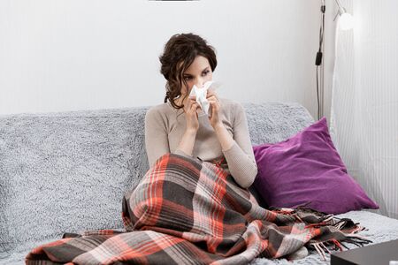 Sick woman suspected of covid-19, being at home in bed, measures the temperature and sneezes. Sick woman sitting under the blanket. Girl is quarantined with suspected coronavirusの写真素材