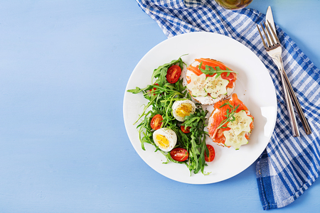 Tasty breakfast. Open sandwiches with salmon, cream cheese and rye bread in a white plate and salad with tomato, egg and arugula. Top view. Flat layの写真素材
