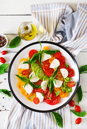 Mozzarella cheese, tomatoes and basil herb leaves in plate on the white wooden table. Caprese salad. Italian food. Top viewの写真素材