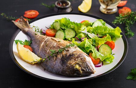 Baked fish dorado with lemon and fresh salad in white plate on dark rustic background. Healthy dinner with fish concept. Dieting and clean eatingの写真素材