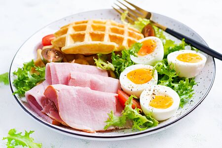 Breakfast with cornmeal waffles, boiled egg, ham and tomato on white background. Appetizers, snack, brunch. Healthy food.の写真素材