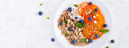 Bowl of flakes with yogurt, blueberryes and persimmon on white wooden table. Fitness food. Overhead,...
