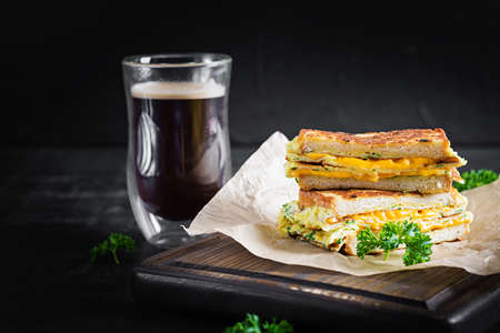 Roasted sandwich cheese and scrambled eggs on paper with coffee glass cup on wooden board. Dark background.の写真素材