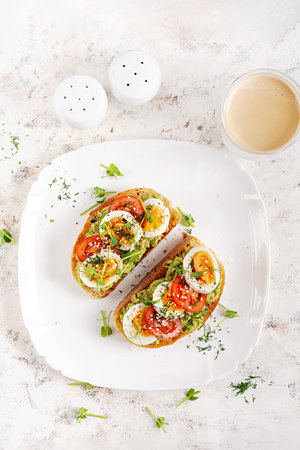 Delicious toast with avocado, boiled egg, tomatoes and microgreen on a white plate. Healthy eating,...