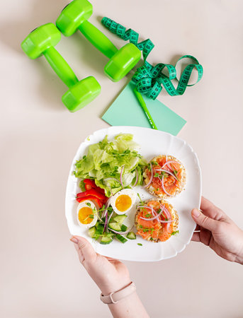 Breakfast with 2 sandwiches with salmon, boiled egg and fresh vegetable salad. The plate is held by female hands, on the table there is a notebook, pen, dumbbells and measuring tape. Flat layの写真素材