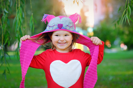 Beautiful laughing little toddler girl in a red coat and colorful knitted hat and scarf playing with yellow maple leaves in a sunny autumn parkの写真素材