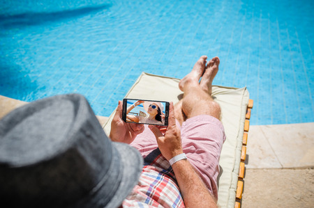 man in a hat lying on a lounger with a smartphone near poolの写真素材