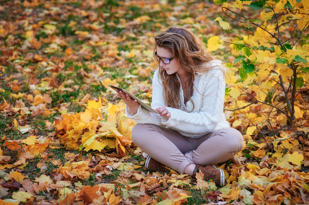 beautiful young woman with a tablet sitting in the autumn park.の写真素材
