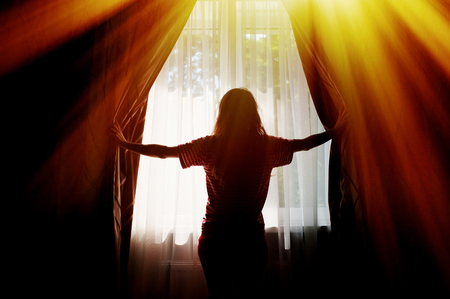 Silhouette of a young woman opens curtains at window.の写真素材