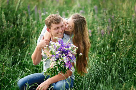 beautiful woman with a bouquet of wild flowers hugging man sitting on the grass in a meadow.の写真素材