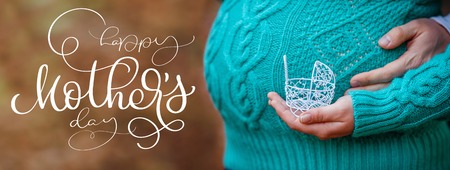 Pregnancy concept: belly with hand holding small toy stroller in front and text Happy mothers day. Calligraphy lettering hand drawの写真素材