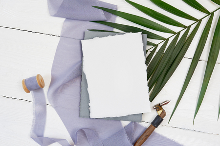 White blank greeting card ribbon on a background of blue fabric with palm leaf and calligraphic pen on a white background. Mockup with envelope and blank card. Flat lay. Top viewの写真素材