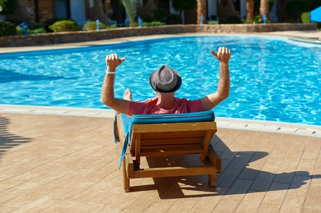 Young man in hat and pink t-shirt lies on louger and relax near swimming pool hotel