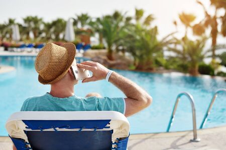 Man in hat using mobile phone on vacation by the pool in hotel, concept of a freelancer working for...