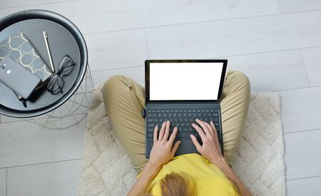 Top view of female freelancer sitting on floor near sofa at home and working on mockup laptop...