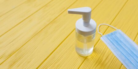 Hand sanitizer bottle and respiratory face mask on yellow wooden background. Stop coronavirus concept banner. Using antibacterial gel for hands during quarantineの写真素材
